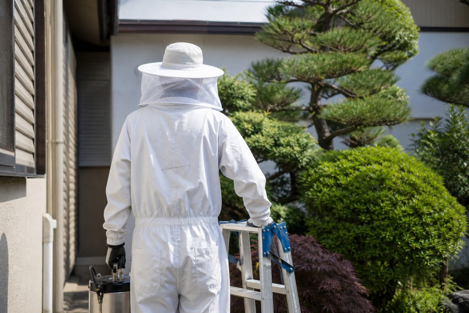 防護服と帽子網を着用し、噴霧器と脚立を持って住宅の庭へ向かう作業員の後ろ姿